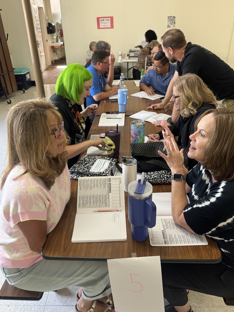 A group of people seated around a table in a classroom with papers, cups, and laptops.