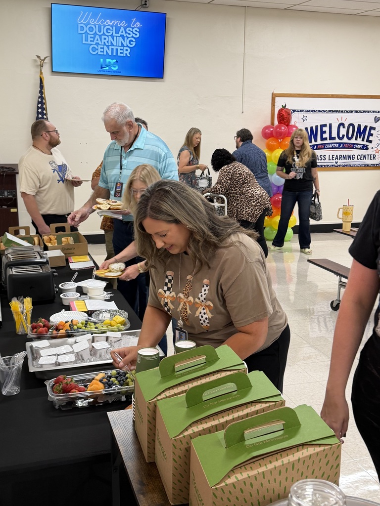 A woman with brown hair serves food from a table. Various people are in the room. A welcome banner and balloons are on the wall.