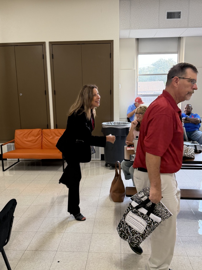 Two people converse in a room with a tiled floor, near a trash can. A woman in black holds a folder. An orange couch and chairs are in the background.