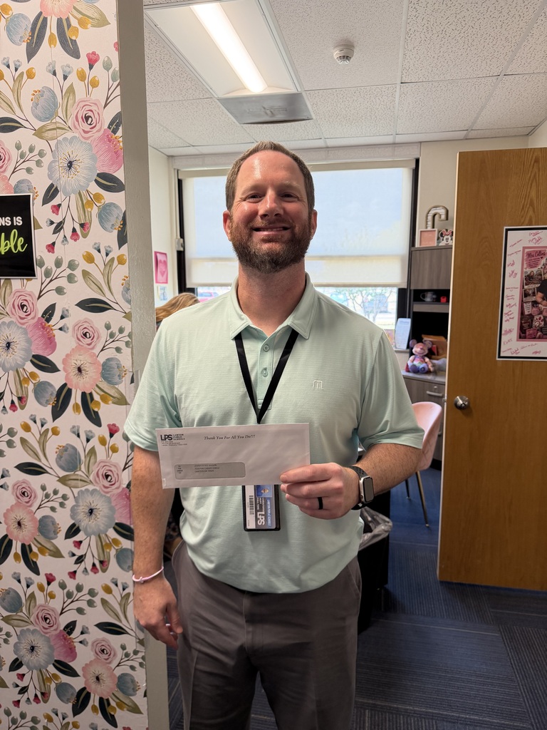A man in a green shirt stands in an office, holding a piece of paper and smiling. Floral wallpaper, a door, and an open window are in the background.