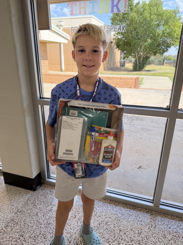 A boy holds a box with various items, smiling. He wears a blue shirt and white shorts. Behind him is a glass door.
