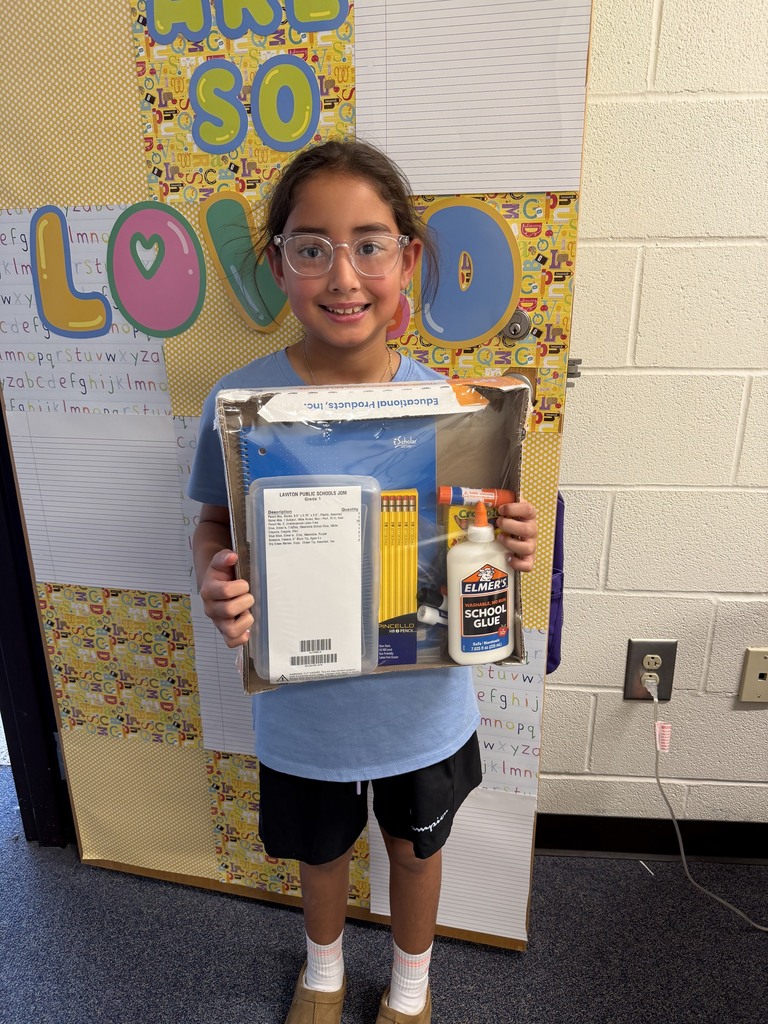 A child in glasses and a blue t-shirt holds a box filled with school supplies. Behind them, a colorful backdrop with words and an electrical outlet is visible.