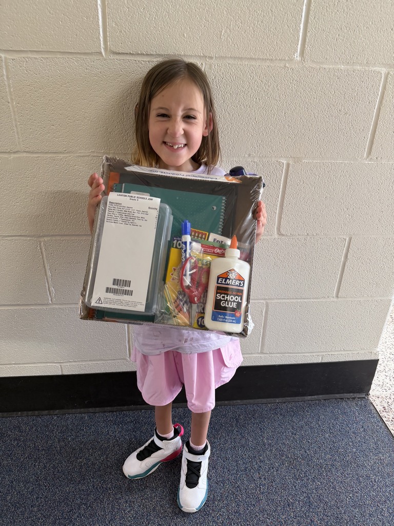 A child with a smiling face holds a box of school supplies in a white room.