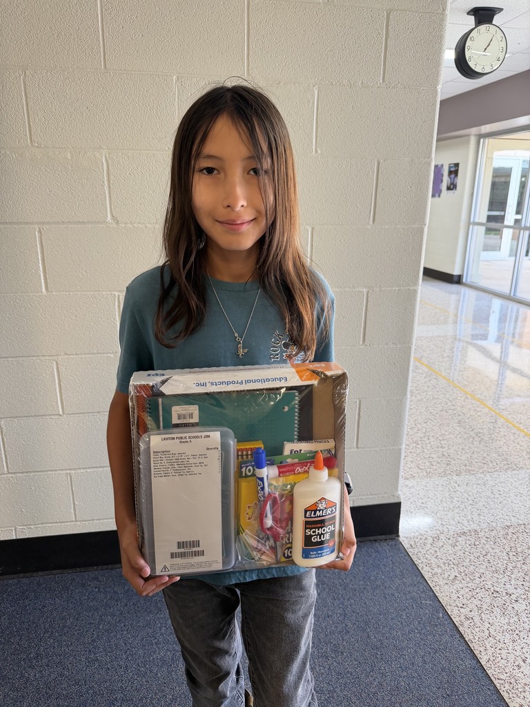 A young girl with long dark hair smiles, holding a box containing school supplies, in a hallway with white brick walls.
