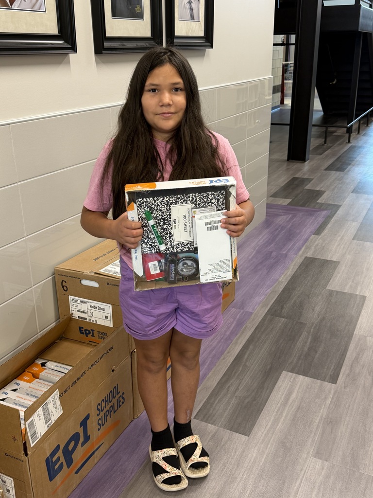 A girl stands holding a box of books, wearing a pink shirt and purple shorts. Behind her, there are boxes and a white wall.