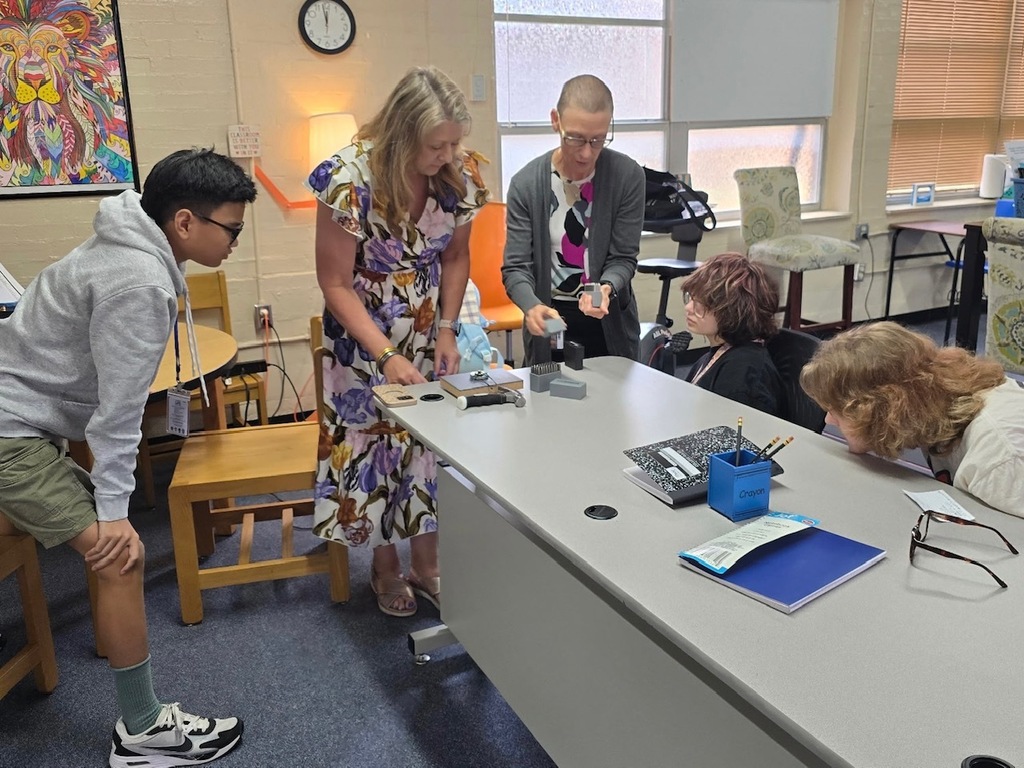 A group of people in a classroom, one leaning over a table, another with a backpack.