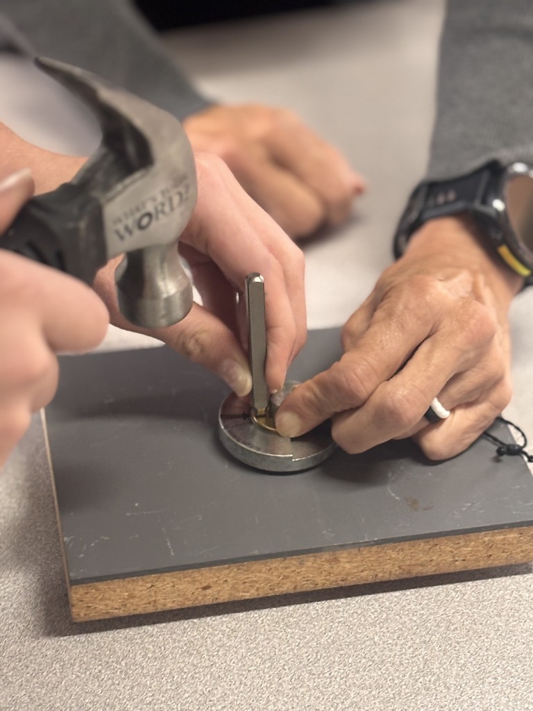 A person holds a hammer over a metal object, which another person is holding on a grey block.