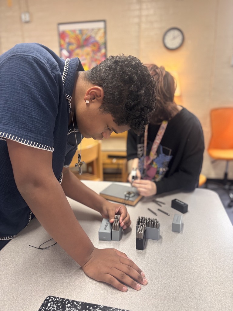 Two students are in a classroom. One is bent over a table with various objects, and the other is seated nearby.