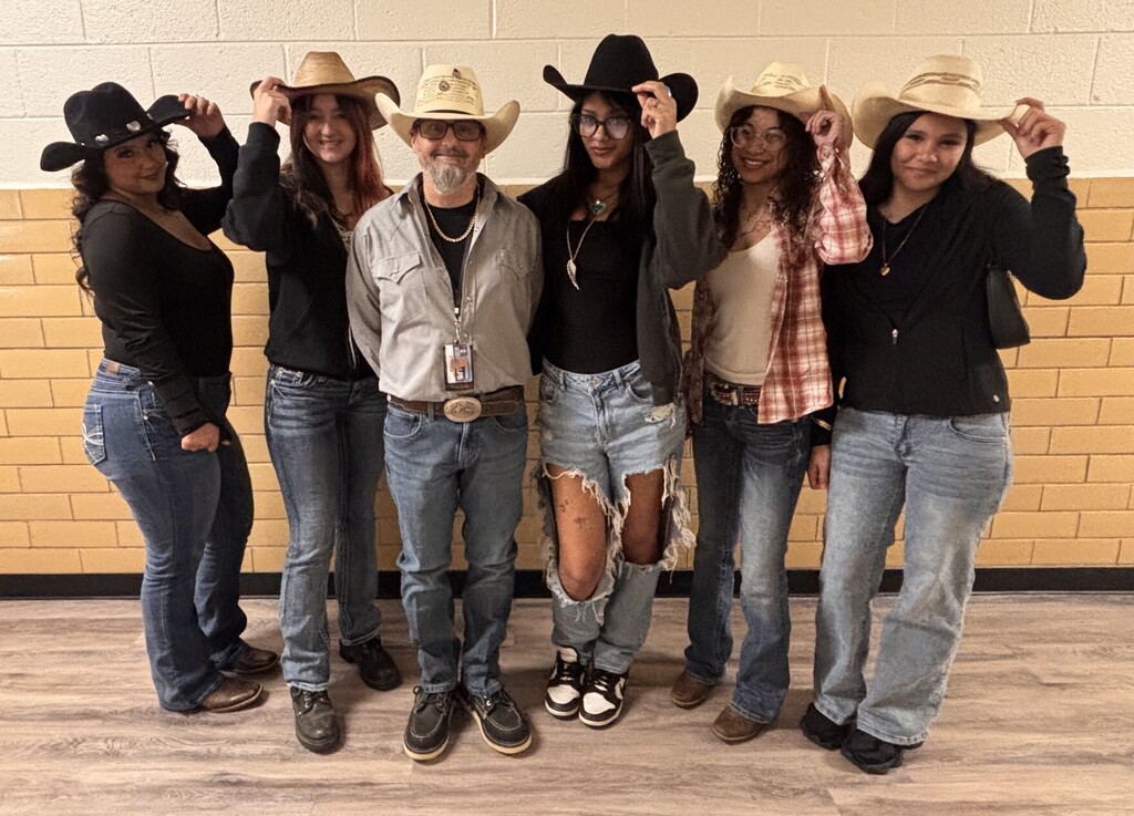 A group of six adults in jeans and cowboy hats, standing together on a wooden floor.