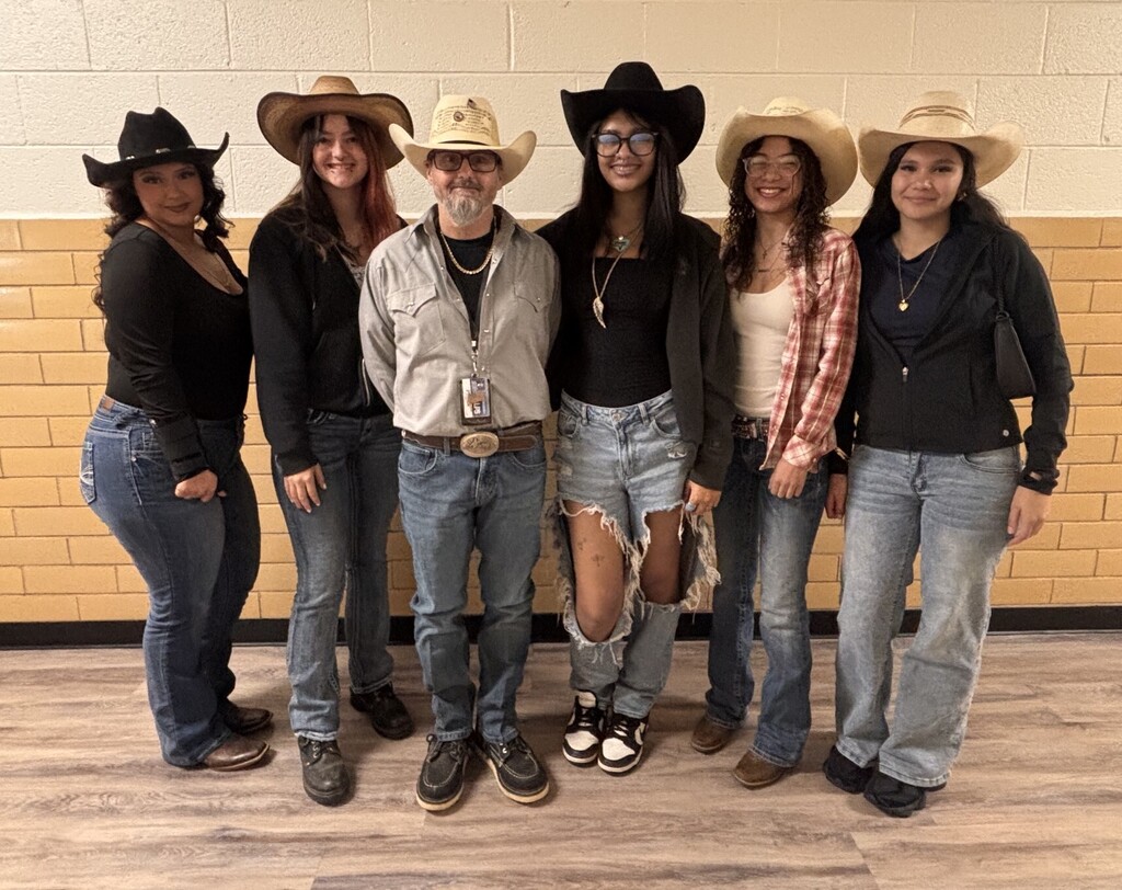Seven people in cowboy hats pose in front of a yellow wall with a wooden floor.
