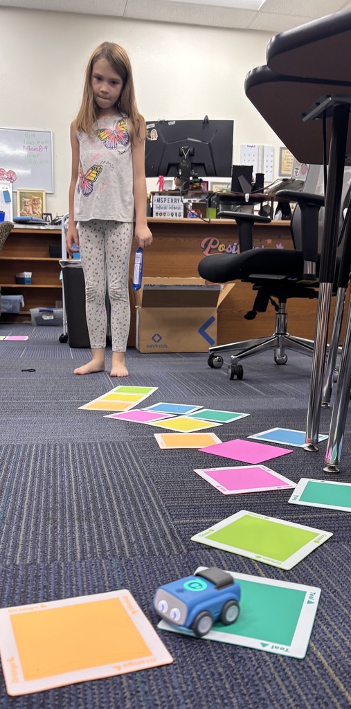 Child standing in room, with multiple colorful squares on floor, one square is blue and a toy car on it.