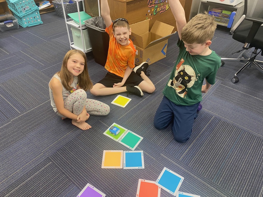 Three children playing with colorful squares on a carpeted floor, one kneeling, another sitting, and the third standing.