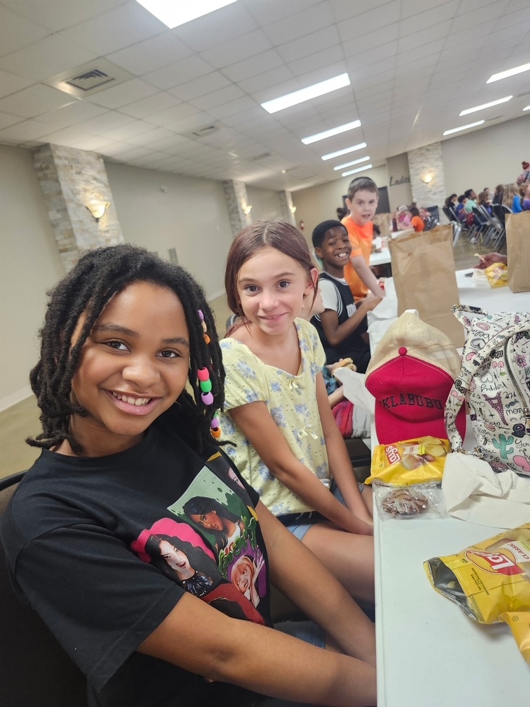 Two girls sitting at a table with food and a bag, smiling, with more people and tables in the background.