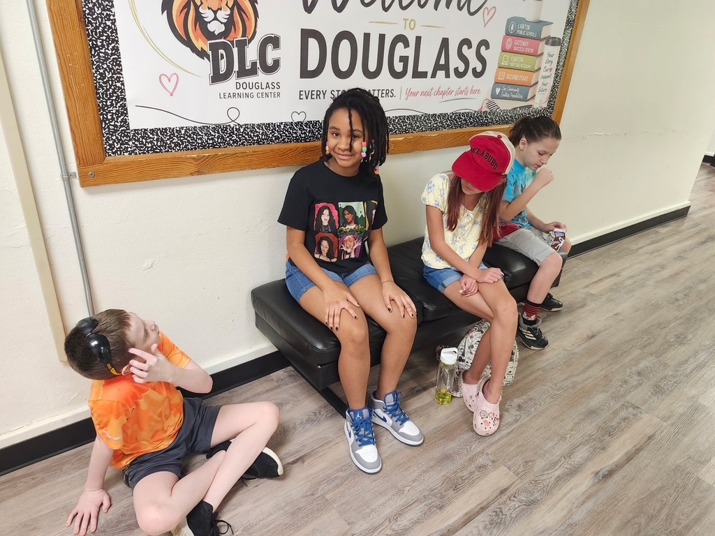 Children seated on a bench in a classroom, one wearing headphones, near a bulletin board.