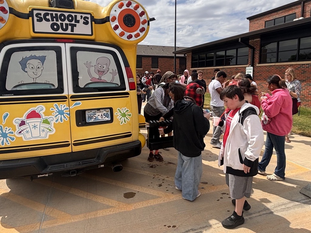 A group of children stand by a yellow school bus with "School's Out" on the back.