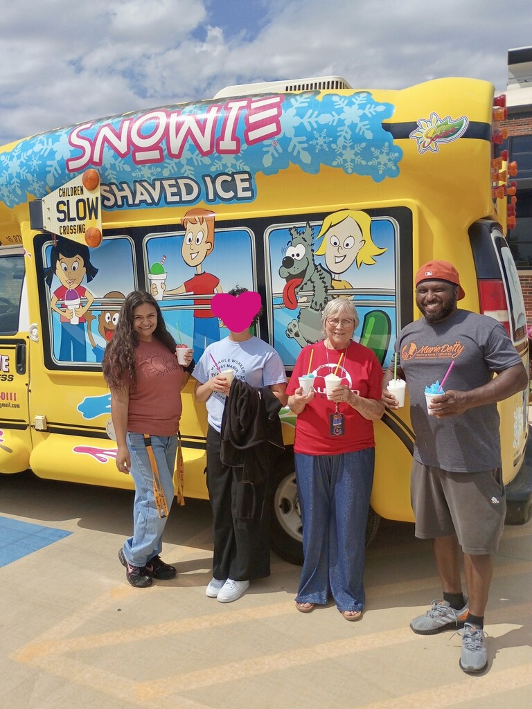 Four people pose in front of a yellow Snopie shaved ice truck with a blue roof. They are holding cups of shaved ice.
