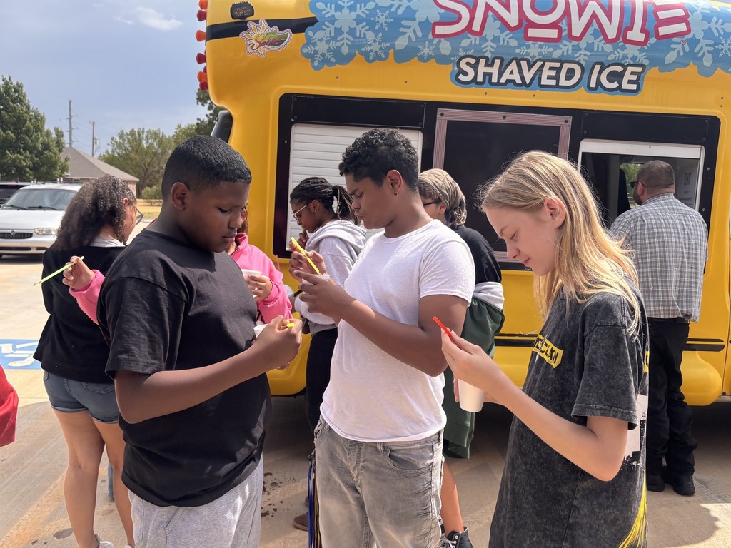 Several people, some eating ice cream, stand before a yellow shaved ice truck on a street.