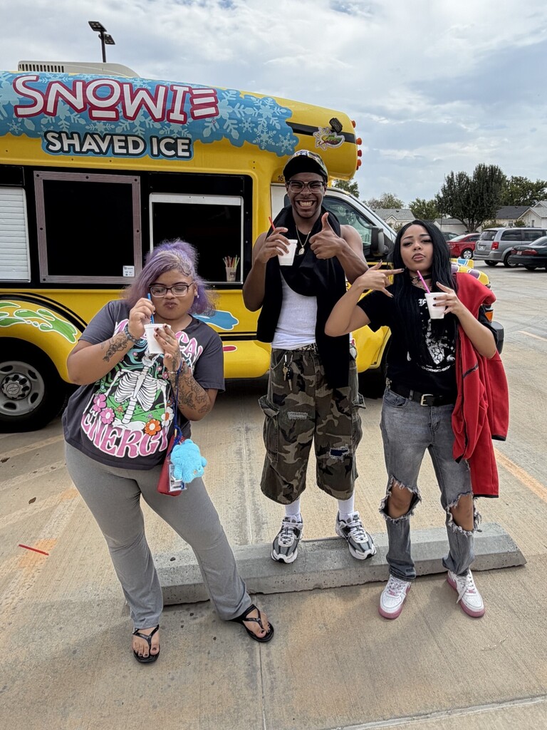 Three individuals in front of a yellow shaved ice truck, each holding cups, smiling and posing.