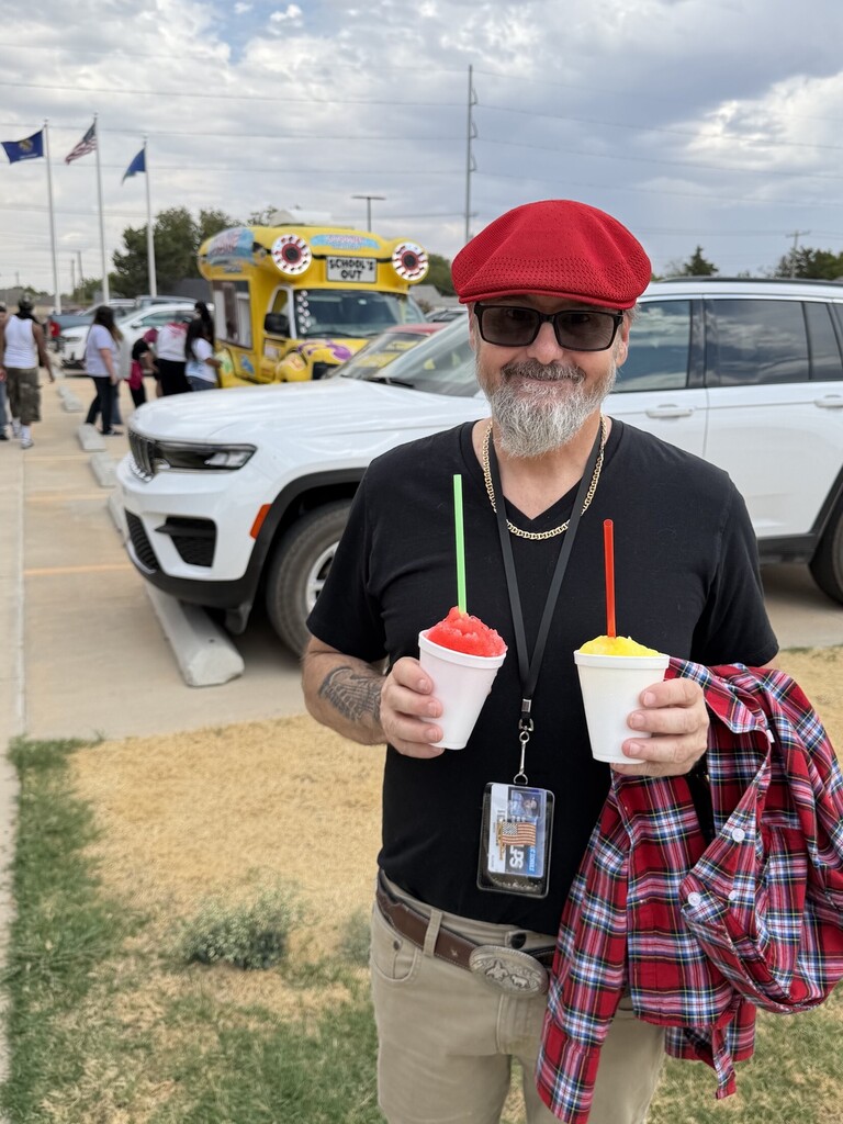 Man in hat holding two snow cones. Background includes a white SUV, a food truck, and people.