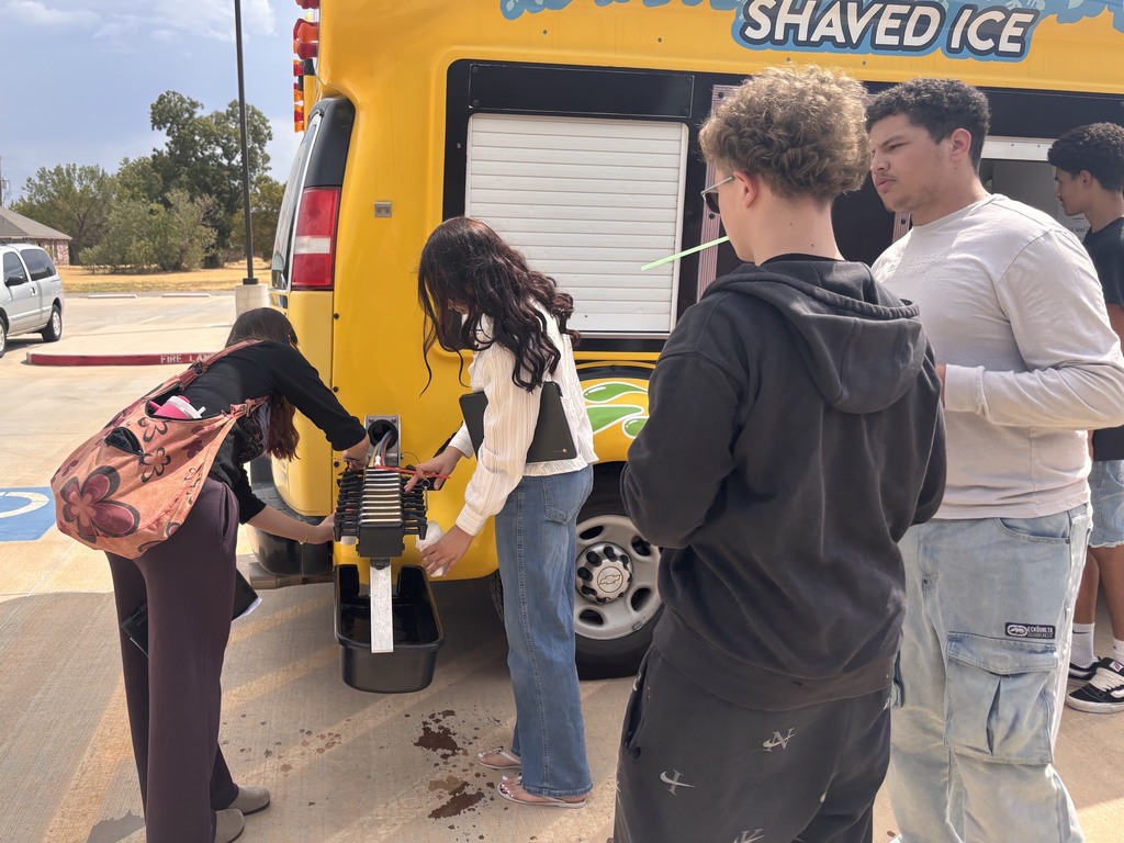 A group of individuals standing around a yellow shaved ice truck. A woman holds a shaved ice machine.