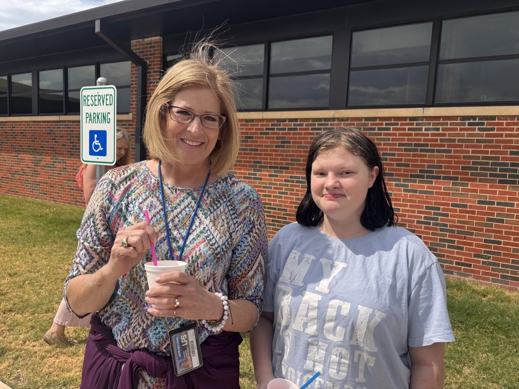 Two women smile outside. One holds a cup, the other wears a gray shirt. Behind them is a brick building.