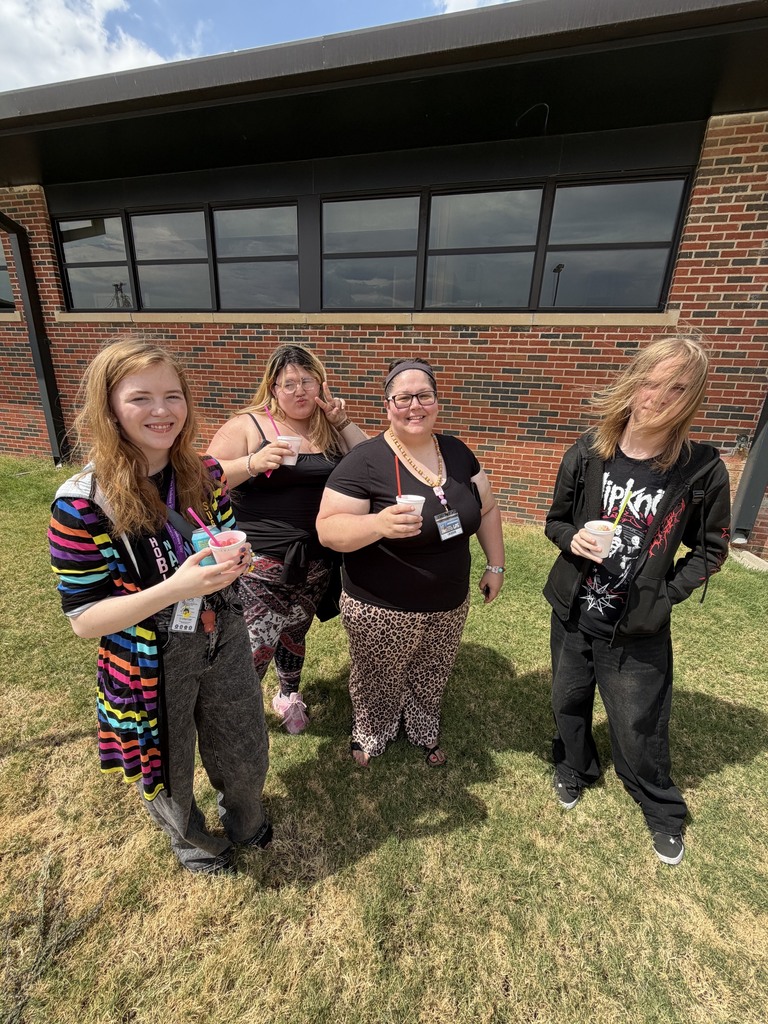 Five women stand in front of a brick building, smiling and holding cups. They wear casual clothes and stand on a lawn.