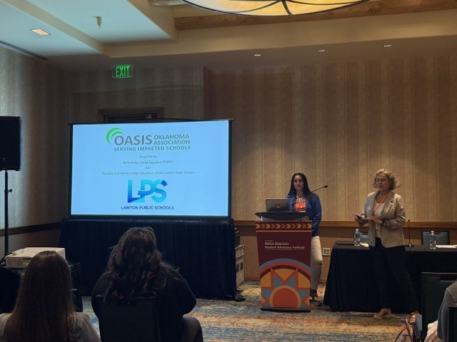 Two women stand at a podium with a screen in the background displaying "OASIS" and "LPS." Audience members are seated.