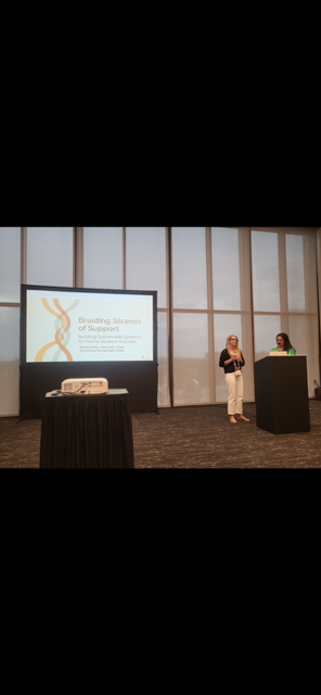 Two women stand in front of a podium with a presentation screen behind them displaying "Braiding Strands of Support."
