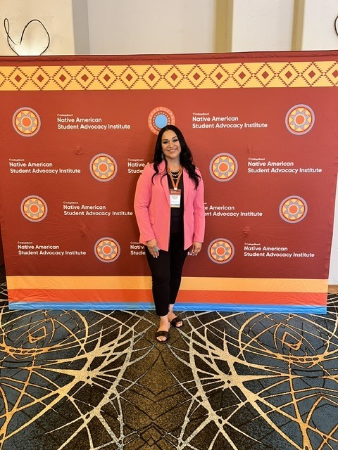 A woman stands in front of a backdrop with a geometric pattern and a title "Native American Student Advocacy Institute".
