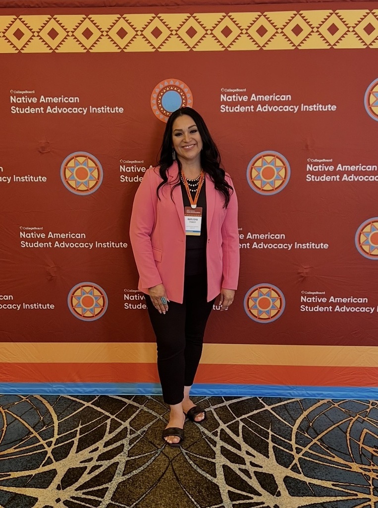 A woman stands in front of a colorful backdrop, featuring a repeating logo, wearing a pink blazer and black pants.