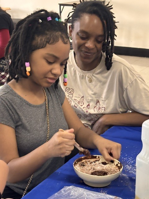 Two women with braided hair sit at a table. One, wearing a gray shirt, stirs chocolate dessert in a bowl.