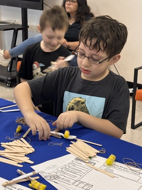 Children are seated at a table, focused on a hands-on activity. One child is using a spoon.