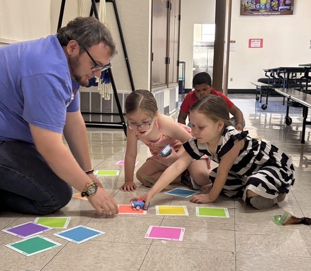 A man helps children with toys on a floor with colored squares. They sit in a classroom area.