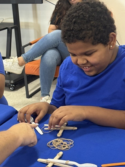 A child sits at a table, focused on assembling a project with wooden sticks and string.