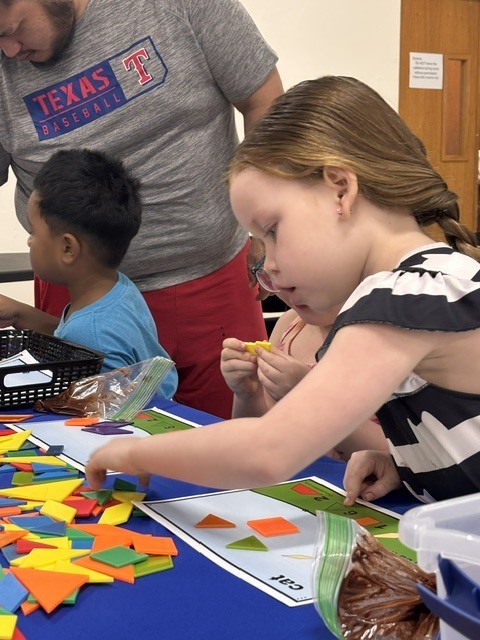 Adults and children gather around a table covered in blue cloth, arranging colorful shapes. A woman looks at the shapes while a child holds a yellow object.