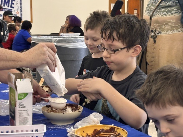 Children at a table, one holding a box, another pouring something into a bowl, with blue tablecloth.