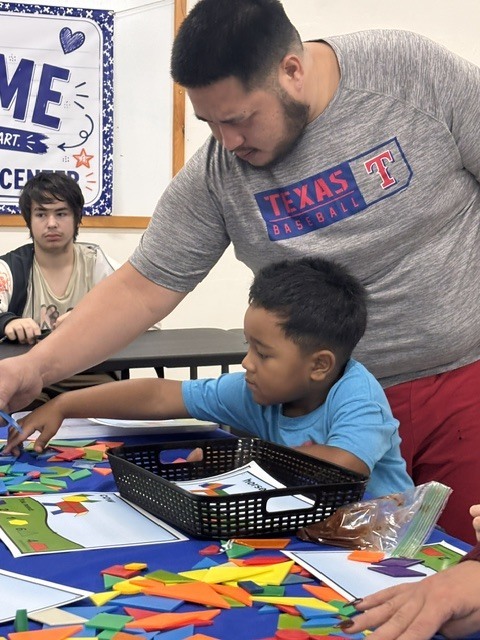 A man assists a child with crafting at a table, surrounded by multicolored papers and a basket.