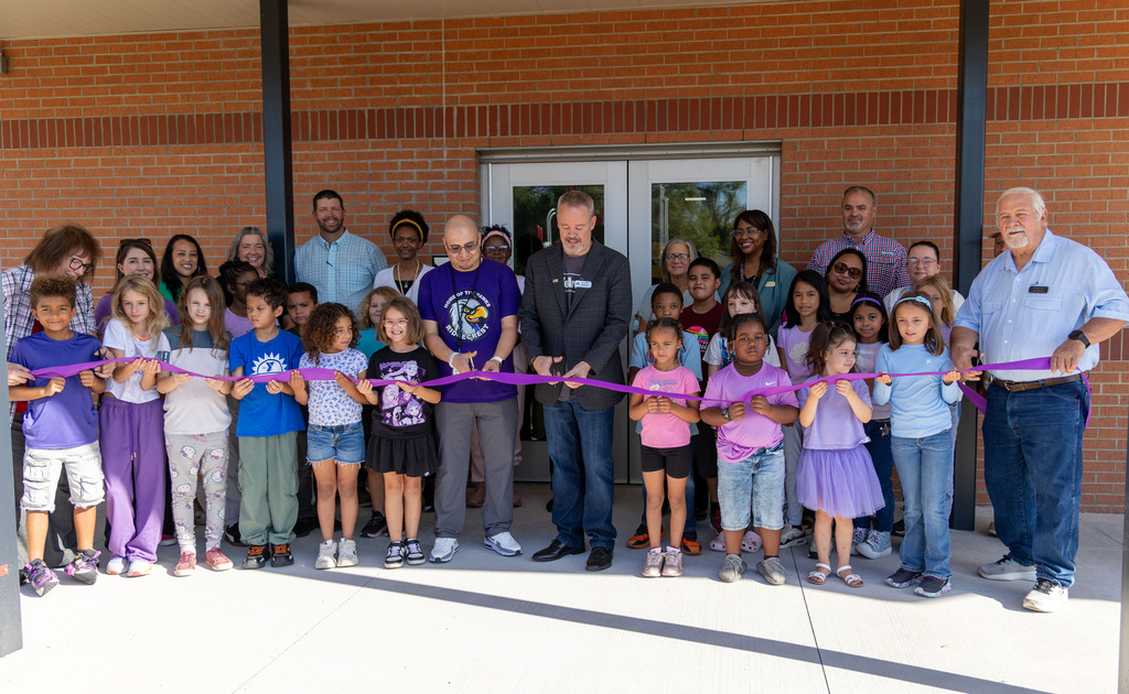 Group of people in front of a brick building, holding a ribbon. Some people wear shoes, others are barefoot.