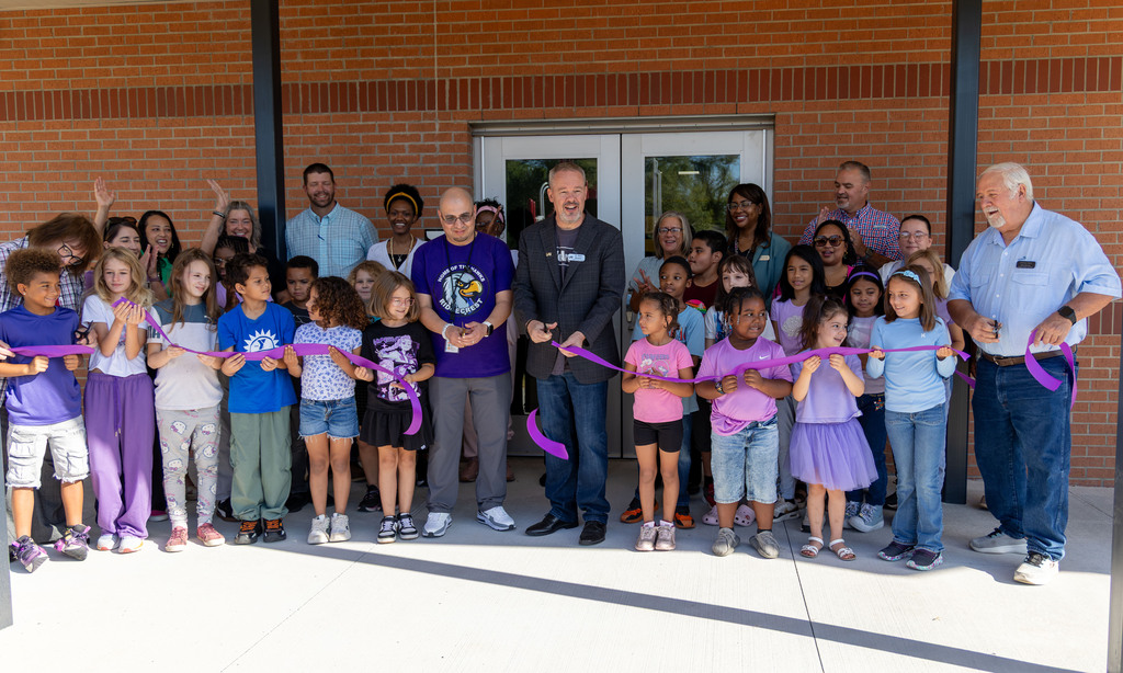 Group of people in front of a brick building, holding a ribbon. Some people wear shoes, others are barefoot.