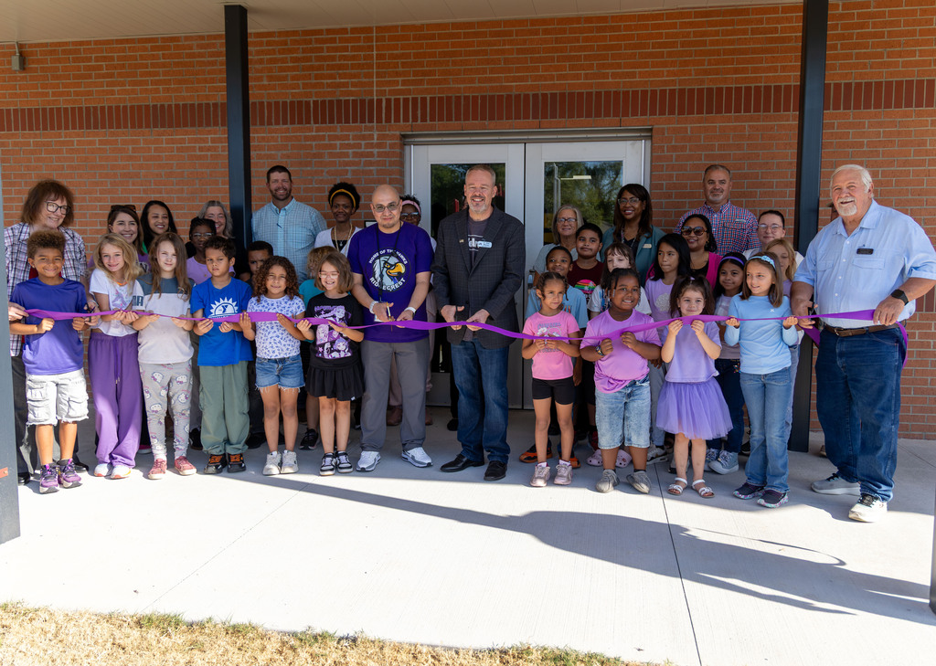 Group of people in front of a brick building, holding a ribbon. Some people wear shoes, others are barefoot.