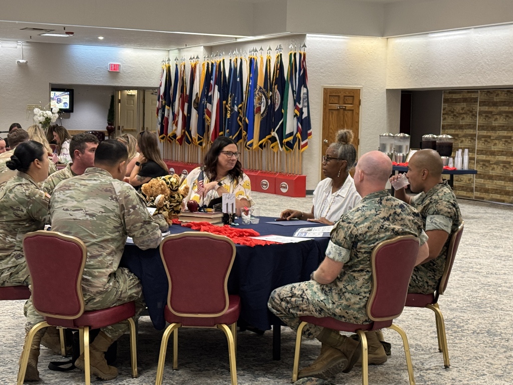People in uniform sit around a table, engaged in conversation. A table displays a blue tablecloth. Flags are visible in the background.