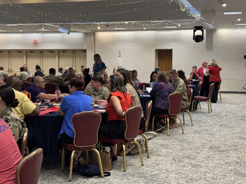 A group of people are seated around tables in a room with a carpeted floor. Some wear military uniforms.