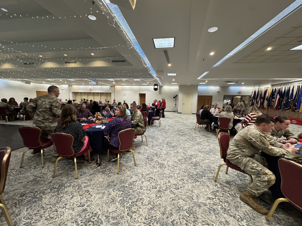 A room with people seated at tables, including individuals in military uniforms, with flags hanging on the wall.