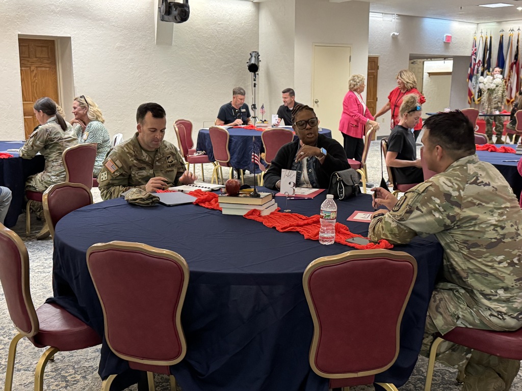 Three people in military uniforms sit around a table with books, papers, and a bottle. Others sit or stand.