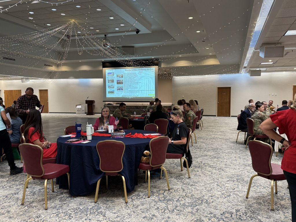 A room with tables and chairs, a projector screen, and people seated or standing around.