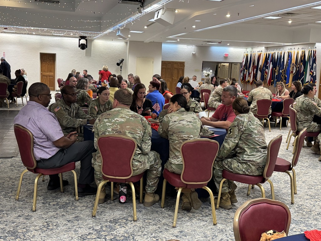 People in uniform and civilian clothes sit at tables inside a banquet hall. Flags line the wall behind them.