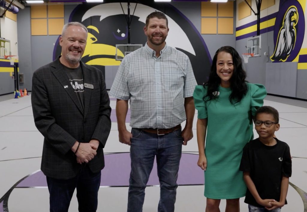 Four people stand in a gymnasium, with a basketball court and school mascot mural in the background.