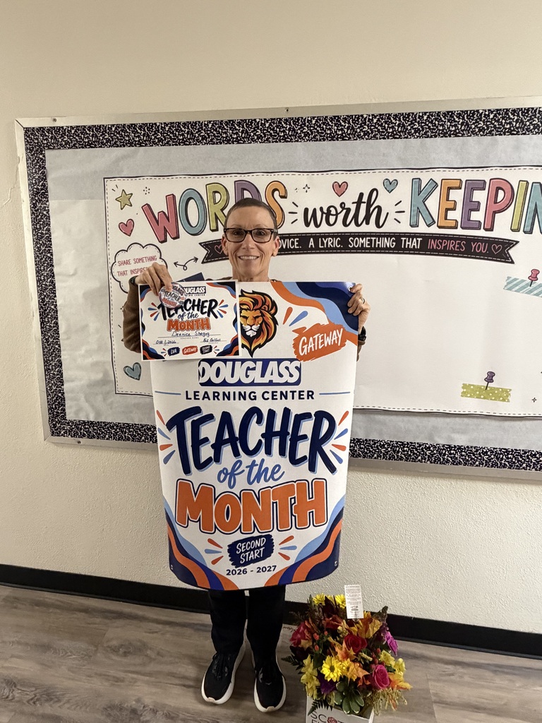 Person standing with a sign reading "Teacher of the Month" in front of a white board.