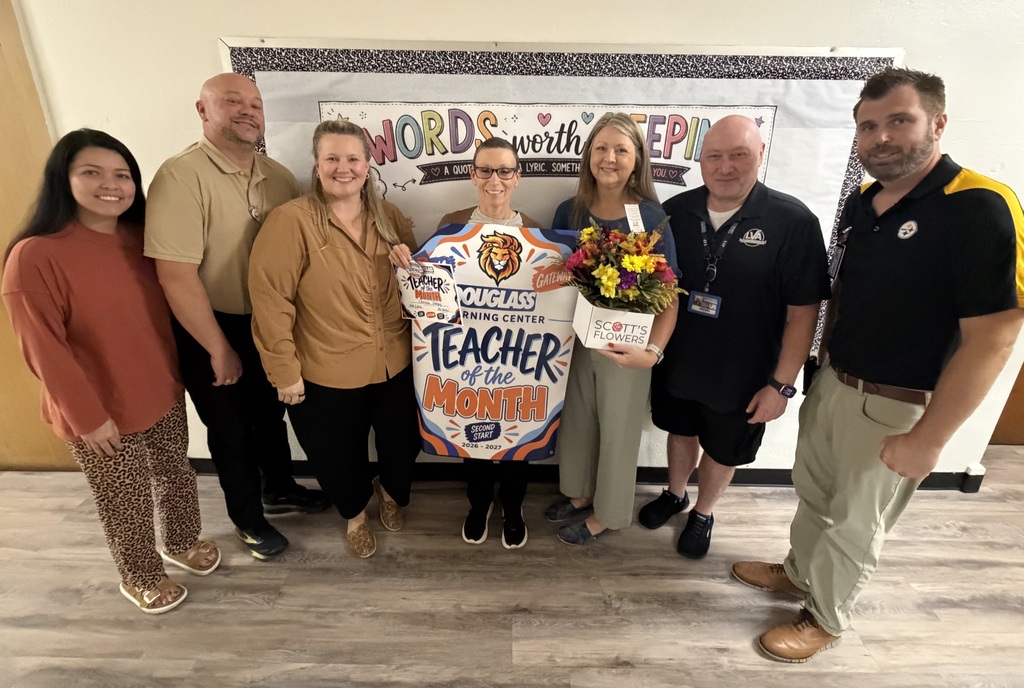 People stand together, smiling, with a banner that reads "Teacher of the Month" One person holds a certificate.