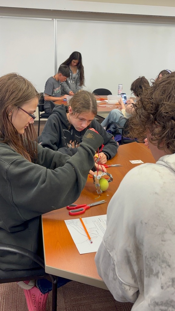 Several students gather around a table. One is holding scissors and appears to be cutting.